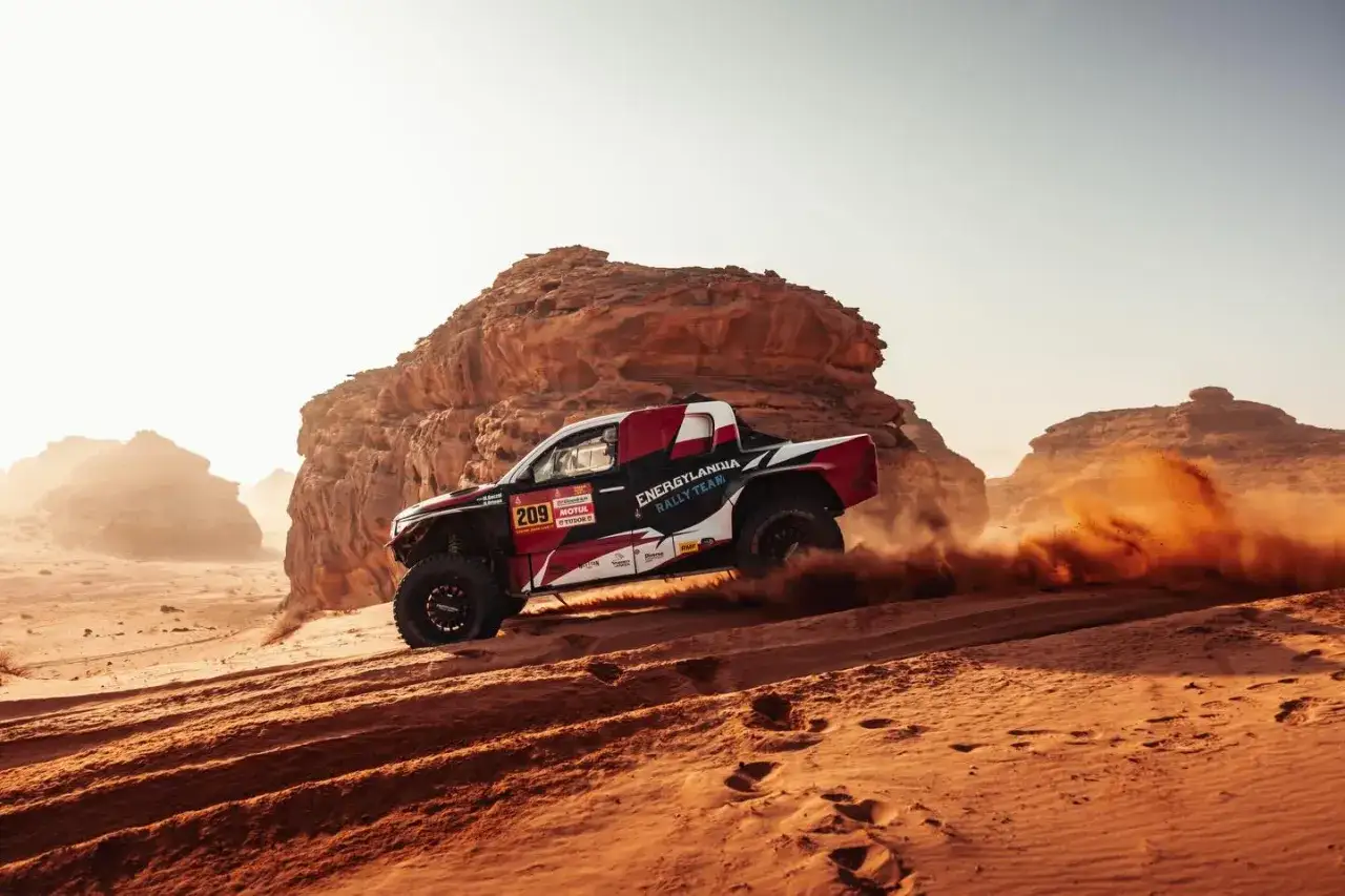 Rajdowy samochód zespołu Energylandia Rally Team pędzi przez pustynię, wzbijając tumany piasku.