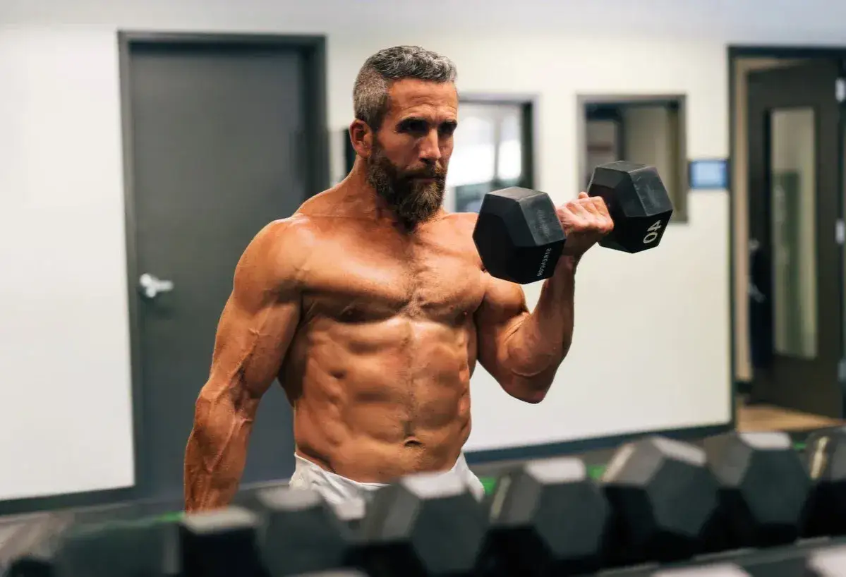 Mann mit Bart hebt Hantel im Fitnessstudio. Seine Muskeln sind definiert, ein Zeichen f&uuml;r Body Recomposition.