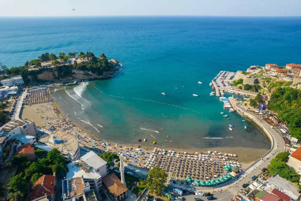 Widok z lotu ptaka na zatokę z plażą pełną leżaków i parasoli. Widać port z łodziami i starą twierdzę. To jedne z głównych ulcinj atrakcje.