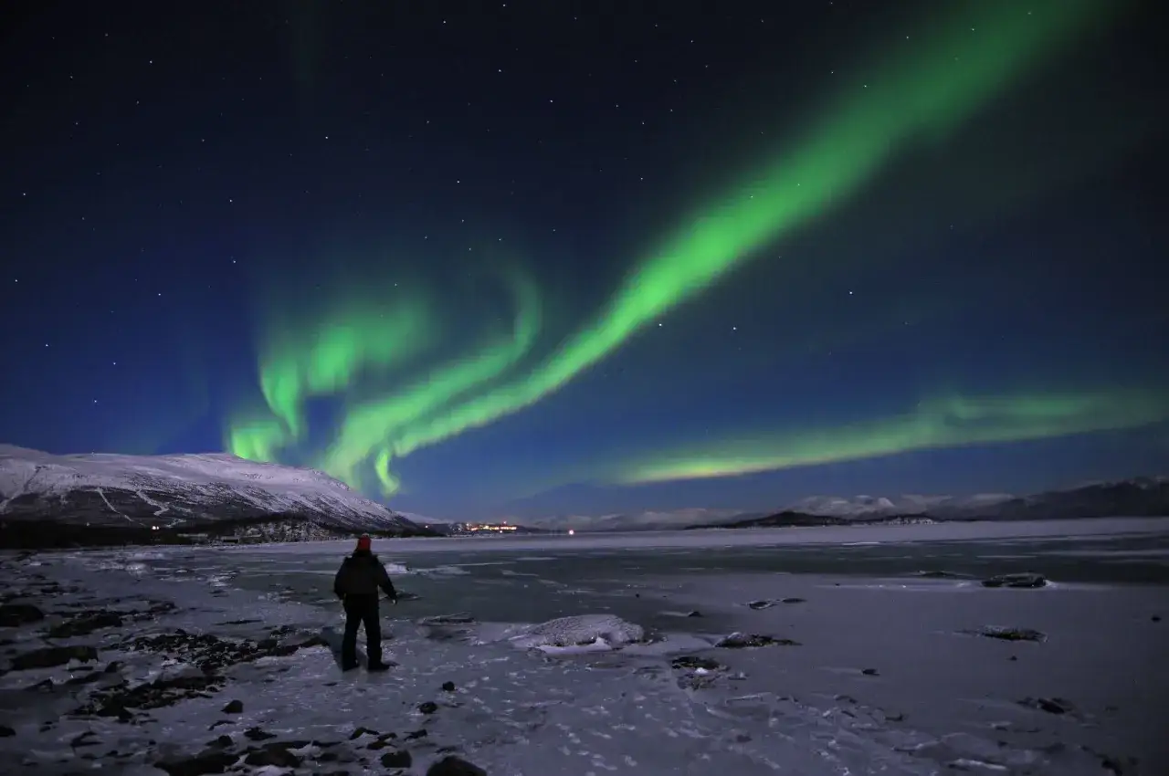 En person st&aring;r p&aring; en isig strand och ser norrsken &ouml;ver ett sn&ouml;t&auml;ckt landskap. En magisk norrsken resa i Sverige.