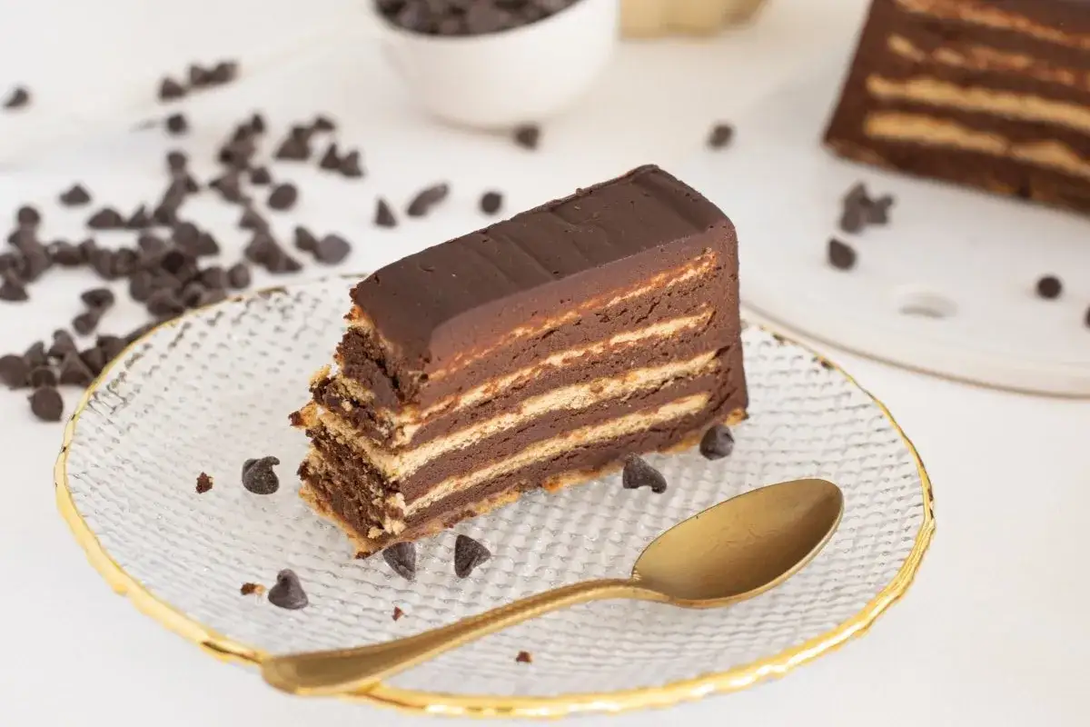 Un trozo de pastel de chocolate con capas, como el que hacía la abuela en España, servido en un plato con chispas de chocolate y una cuchara dorada.