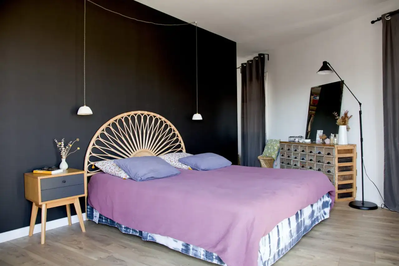 Chambre avec mur noir mat contrastant avec des rideaux sombres. Un lit violet est orné d'un tête de lit en rotin.
