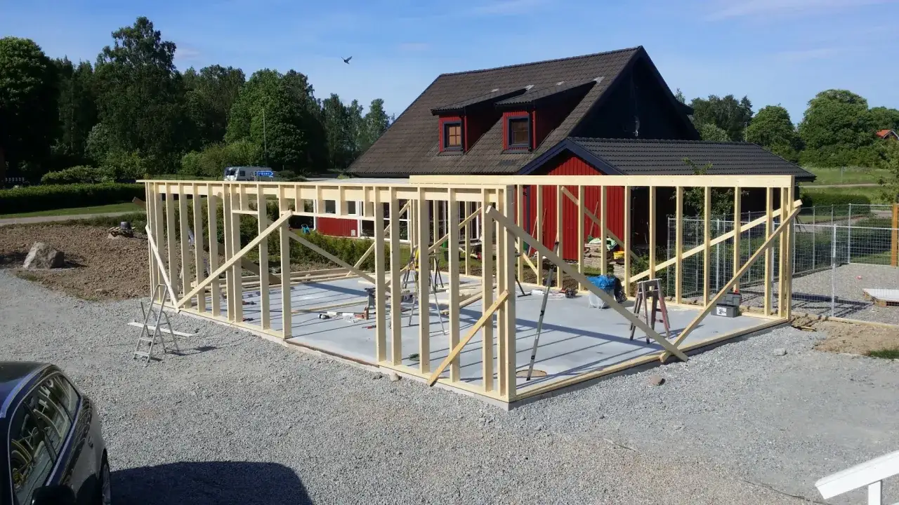Grundläggande stomme för att bygga garage på en betongplatta. Träbjälkar reses mot ett rött hus i bakgrunden.