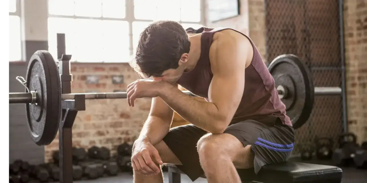 Erschöpfter Mann sitzt auf einer Bank im Fitnessstudio, die Stirn gerunzelt, vielleicht ein Fehler beim Muskelaufbau. Hanteln und Gewichte im Hintergrund.