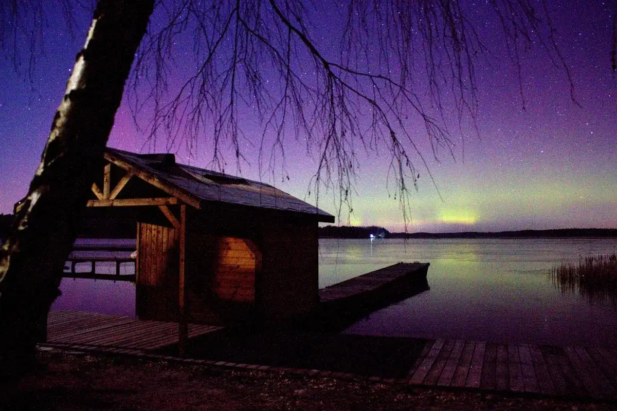 Drewniana chata nad jeziorem, nad którą rozciąga się fioletowo-zielona zorza polarna.