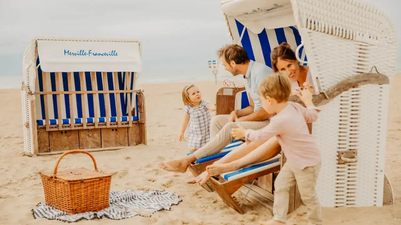 Famille profitant d'une **vacance scolaire en Normandie** à Merville-Franceville. Ils sont assis dans une cabine de plage, avec un panier de pique-nique à proximité.