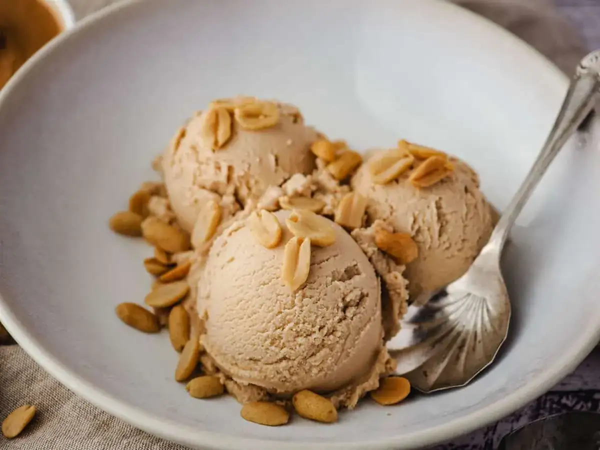 Delicioso helado de crema de cacahuete servido en un cuenco, adornado con cacahuetes tostados. Una cucharilla plateada espera para disfrutar de este postre.
