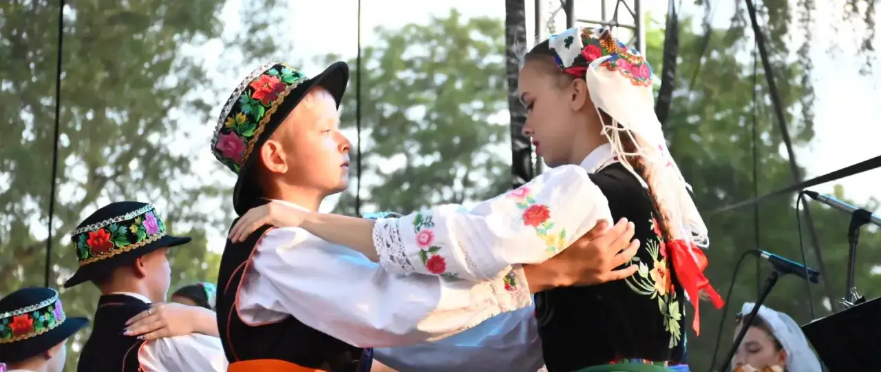 Chłopiec i dziewczyna w tradycyjnych strojach tańczą polkę. Ich stroje zdobią kolorowe hafty i kwiaty.