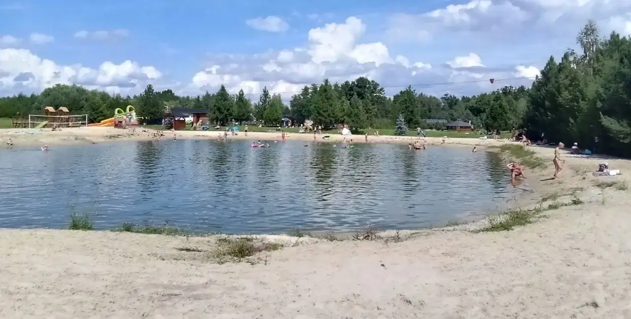 Sielska pogoda sprzyja plażowaniu i kąpielom w jeziorze. Ludzie odpoczywają na piasku, kąpią się i bawią w wodzie.