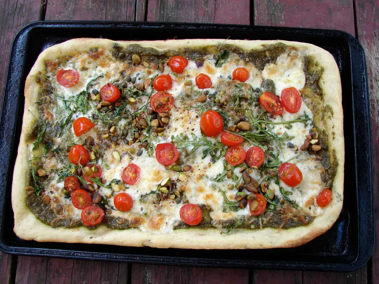 Prostokątna pizza z zielonym pesto, pomidorkami koktajlowymi, pistacjami i rukolą. Idealna na letnie popołudnie, smakuje jak prawdziwa **spinata**!