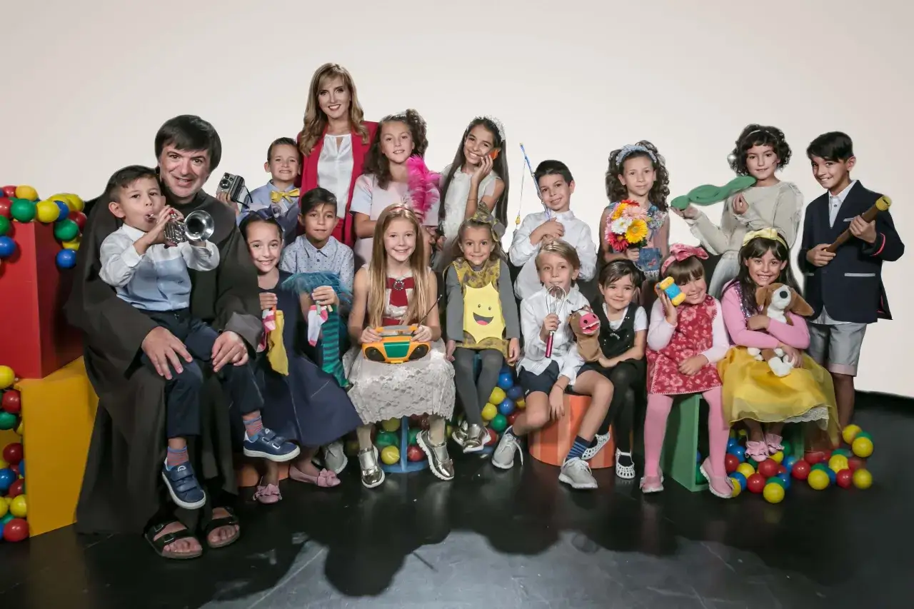 Un gruppo di bambini festosi, alcuni con strumenti musicali e giocattoli, posa con un uomo e una donna, evocando le 25 canzoni pi&ugrave; famose dello Zecchino d'Oro.