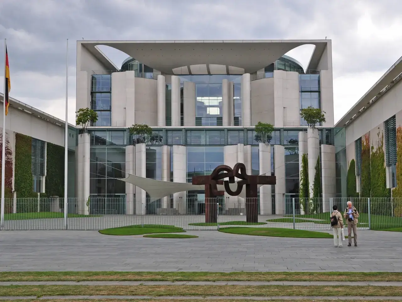 Das Bundeskanzleramt in Berlin, wo der Bundeskanzler wohnt, mit zwei Besuchern vor der modernen Architektur und einer Skulptur.