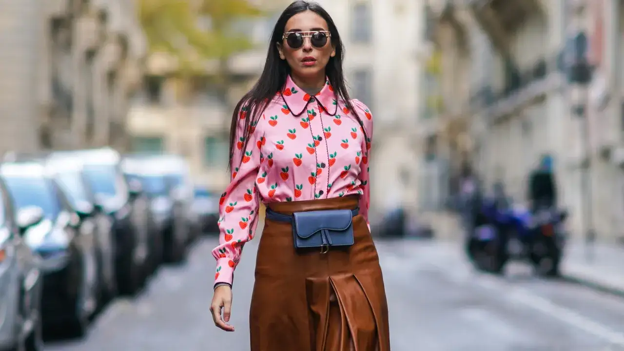 Kobieta w różowej koszuli w truskawki i brązowej spódnicy, z torebką na biodrach, prezentuje styl preppy.