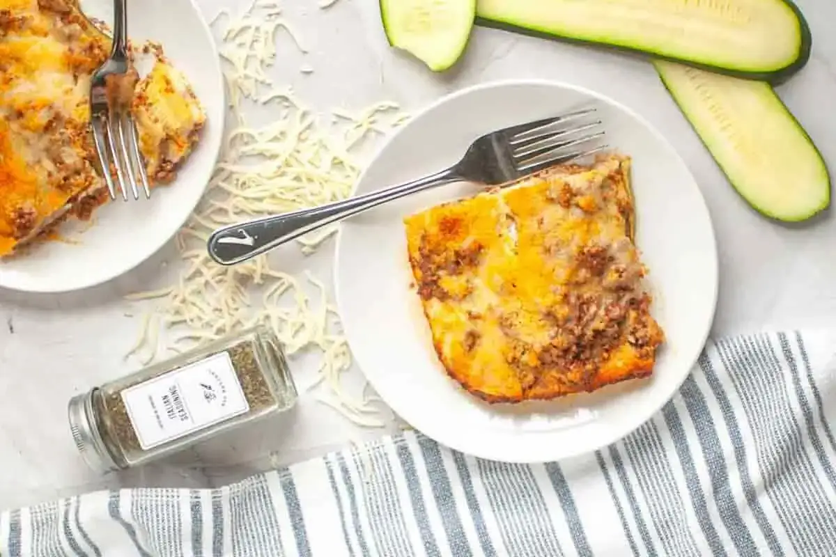 Low carb Lasagne mit Hackfleisch und K&auml;se, serviert auf einem Teller mit Zucchini-Scheiben und italienischen Gew&uuml;rzen.
