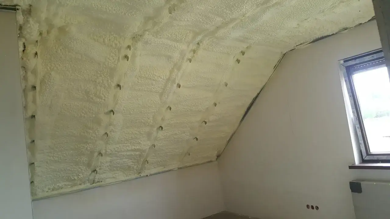 Pianka do izolacji termicznej pokrywa skośny sufit poddasza, tworząc jednolitą, puszystą warstwę. Widać metalowe elementy mocujące.