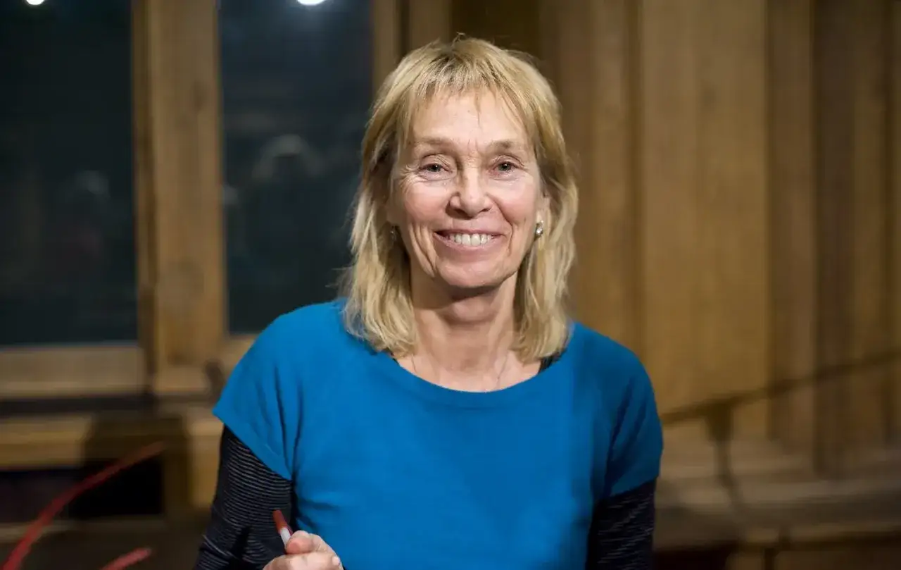 Uśmiechnięta Małgorzata Braunek w niebieskiej bluzce, z blond włosami i wisiorkiem na szyi, stoi na tle drewnianej ściany.