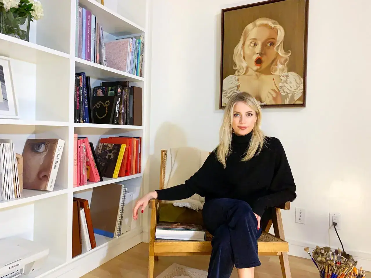 Anna Weyant, artist, sits in a chair near a bookshelf and a painting. Larry Gagosian might appreciate this scene.
