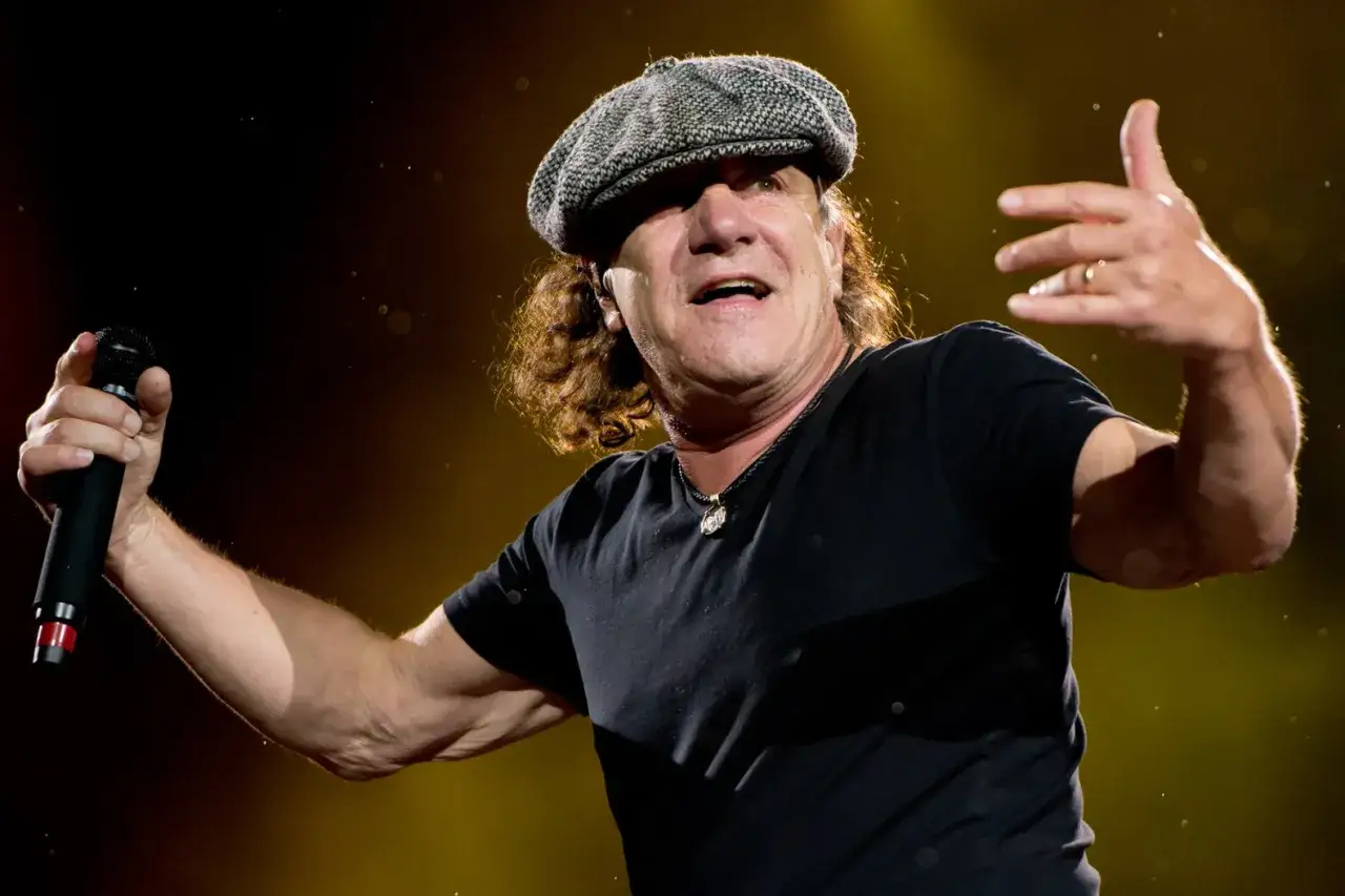Brian Johnson w czapce i czarnej koszulce śpiewa do mikrofonu, gestykulując.