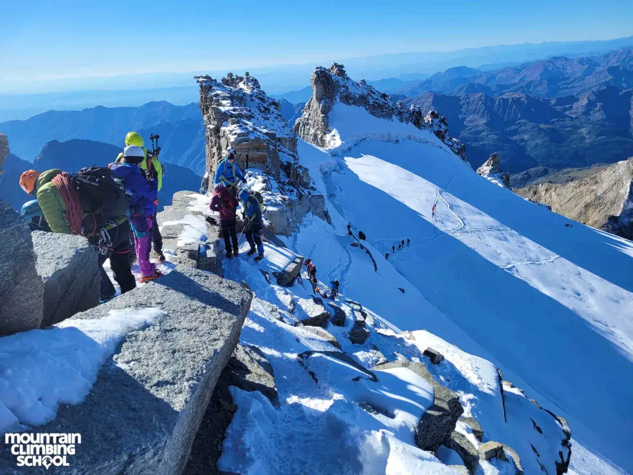 Grupa alpinistów na skalistym, ośnieżonym grzbiecie podczas wspinaczki na **Mont Blanc**. W tle rozległe pasma górskie.