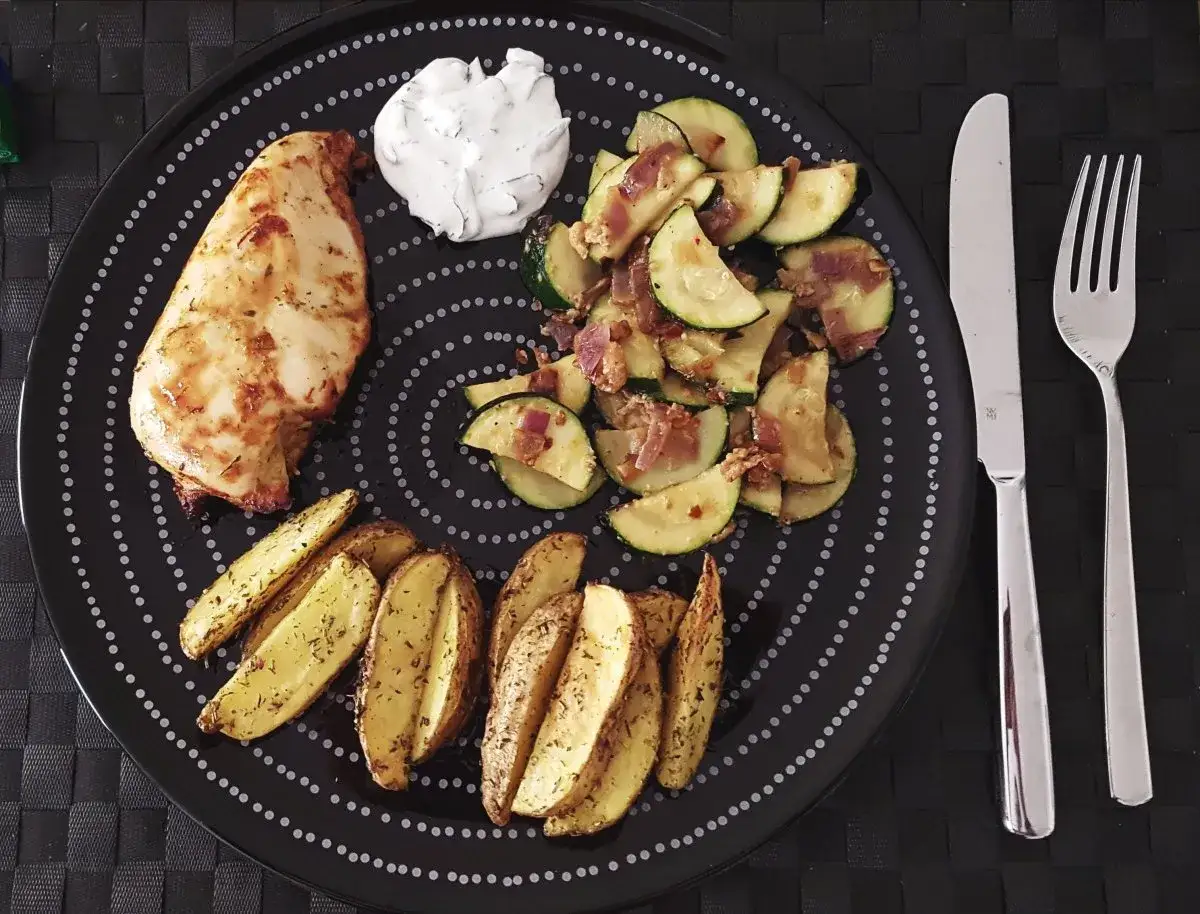 Ein Teller mit Hähnchenbrust, Kartoffelspalten und Zucchinigemüse, perfekt für Airfryer Rezepte. Dazu ein Klecks Dip.