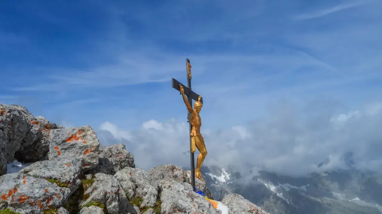 Goldenes Kreuz auf felsigem Gipfel, mit Blick auf Wolken und Berge. Ein Symbol der Hoffnung am hocheck Watzmann.
