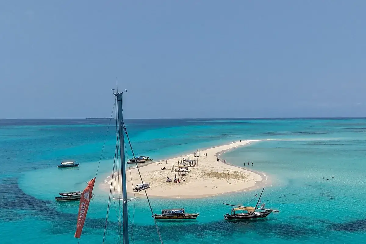 Piaskowa łacha na Zanzibarze, otoczona turkusową wodą. Ludzie spacerują po plaży, a łodzie kołyszą się na falach.