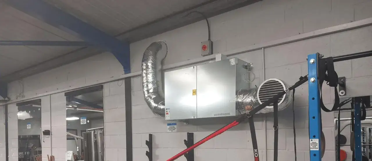 Extractor de aire para habitaci&oacute;n de gimnasio, con conductos plateados y un panel met&aacute;lico en la pared de ladrillos.