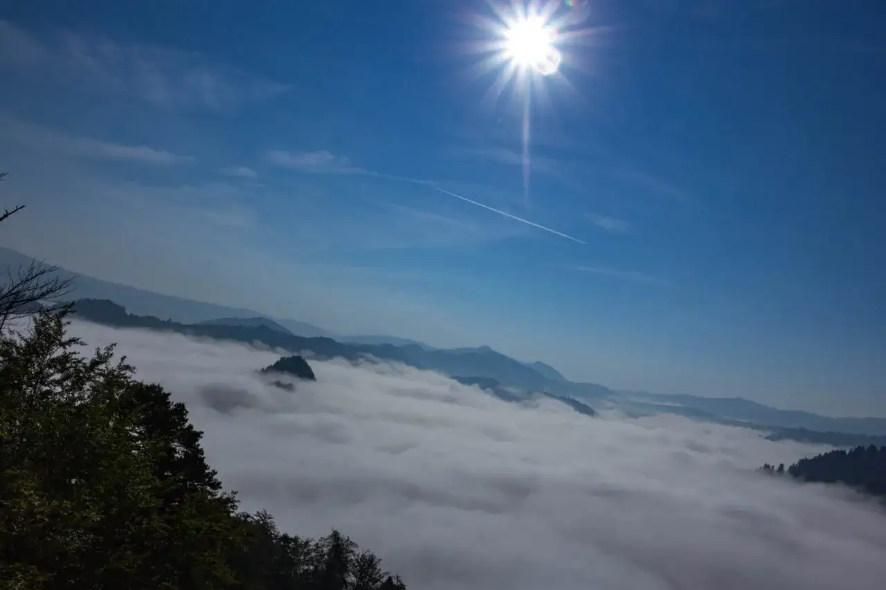 Słońce tworzy efekt lens flare na błękitnym niebie nad morzem mgły spowijającym góry.