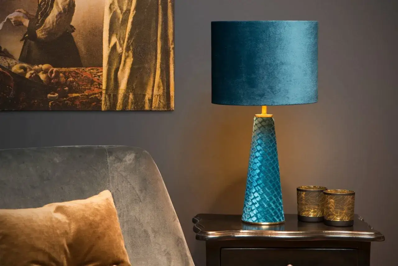 Elegante lámpara turquesa con pantalla de terciopelo azul, junto a un sillón y dos portavelas dorados.