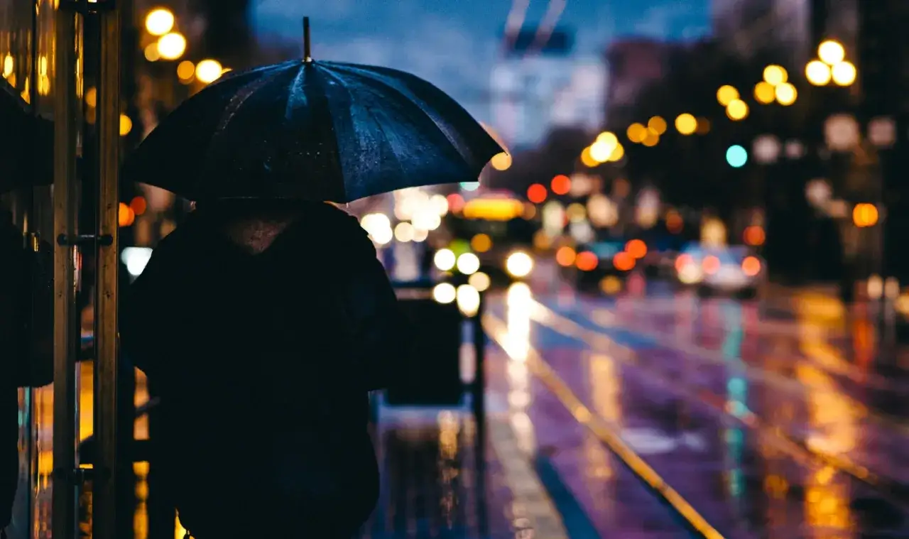 Une personne sous un parapluie, attendant dans la nuit pluvieuse. Les lumi&egrave;res de la ville se refl&egrave;tent sur la chauss&eacute;e mouill&eacute;e, cr&eacute;ant de belles photos sous la pluie.