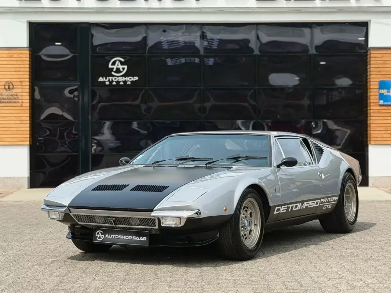 Srebrny De Tomaso Pantera GTS z czarną maską i pasem na boku, zaparkowany przed budynkiem z czarnymi, błyszczącymi drzwiami garażowymi.