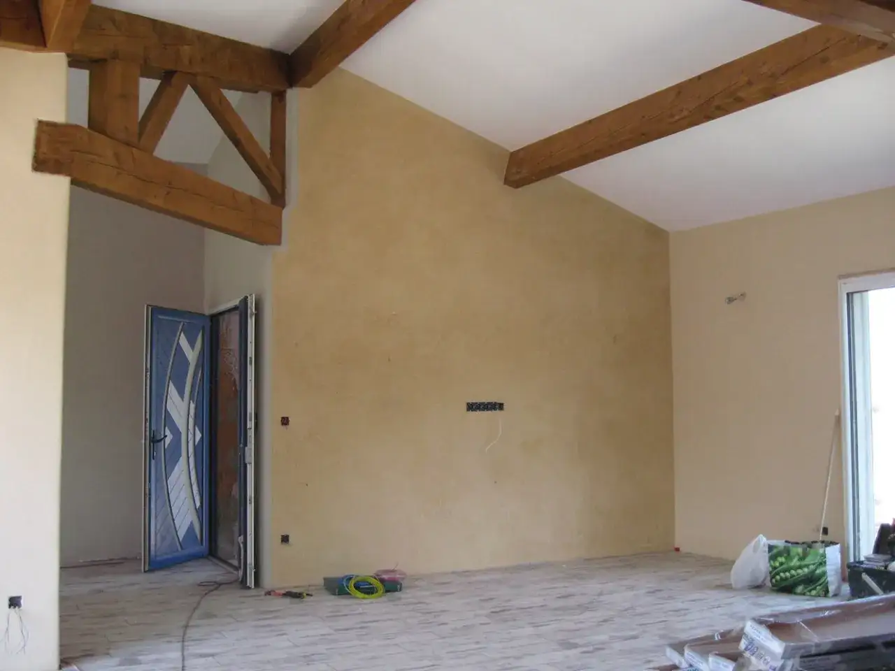 Un grand mur à la chaux dans une pièce en rénovation, avec des poutres apparentes au plafond et une porte bleue ouverte.