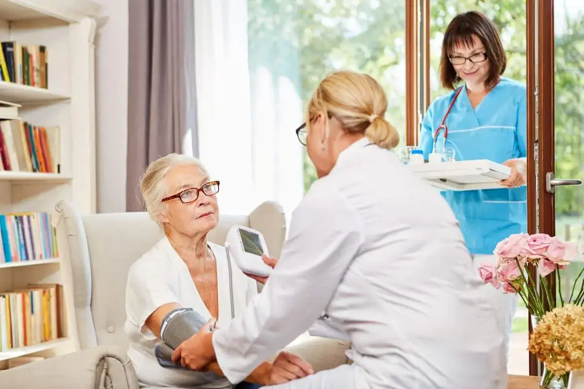 Opiekunka mierzy ciśnienie starszej kobiecie, która może mieć objawy alzheimera. Druga pielęgniarka niesie tacę z lekami.