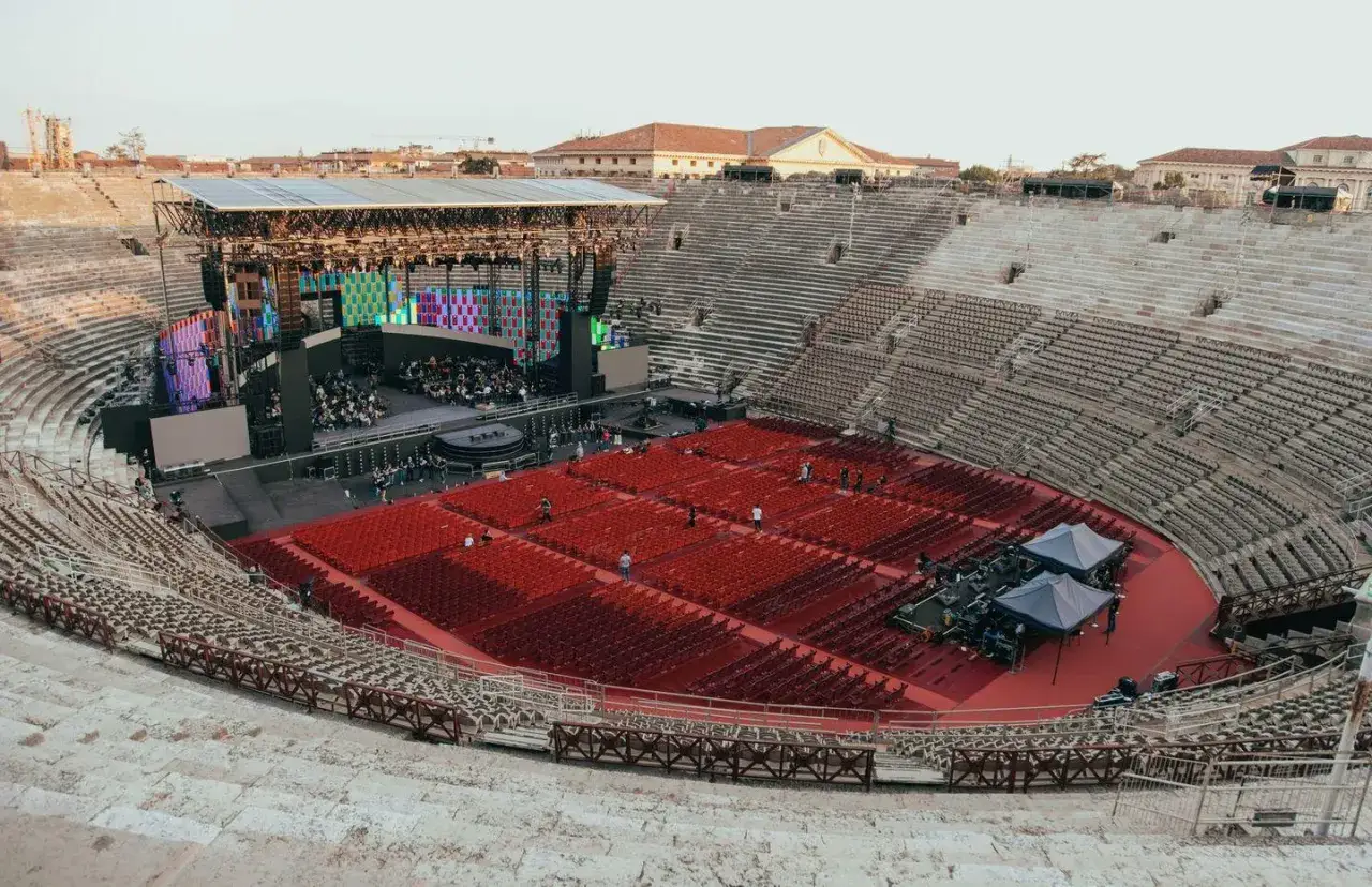 Arena con palco imponente e file di sedie rosse pronte per il pubblico. L'atmosfera è carica di attesa, quasi un preludio a un grande evento, un vero e proprio
