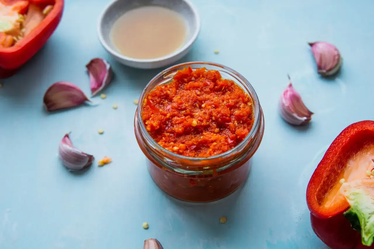 Domowy sos sriracha z papryczek chilli i czosnku w słoiku, obok przekrojona papryka i ząbki czosnku.