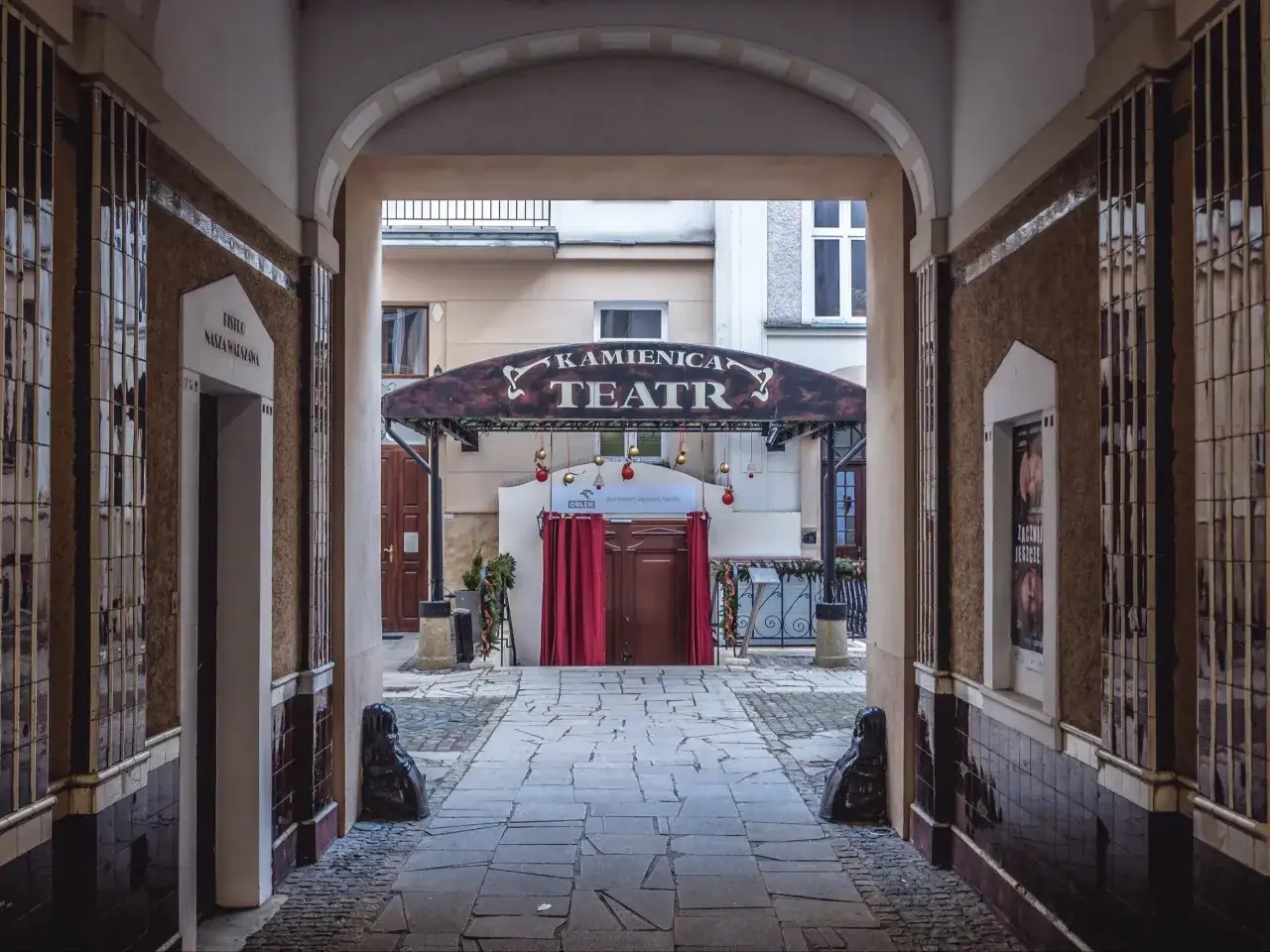 Kamienica Teatr, miejsce stworzone przez założyciela teatru kamienica, zaprasza na spektakl. Czerwone zasłony i świąteczne ozdoby tworzą magiczną atmosferę.