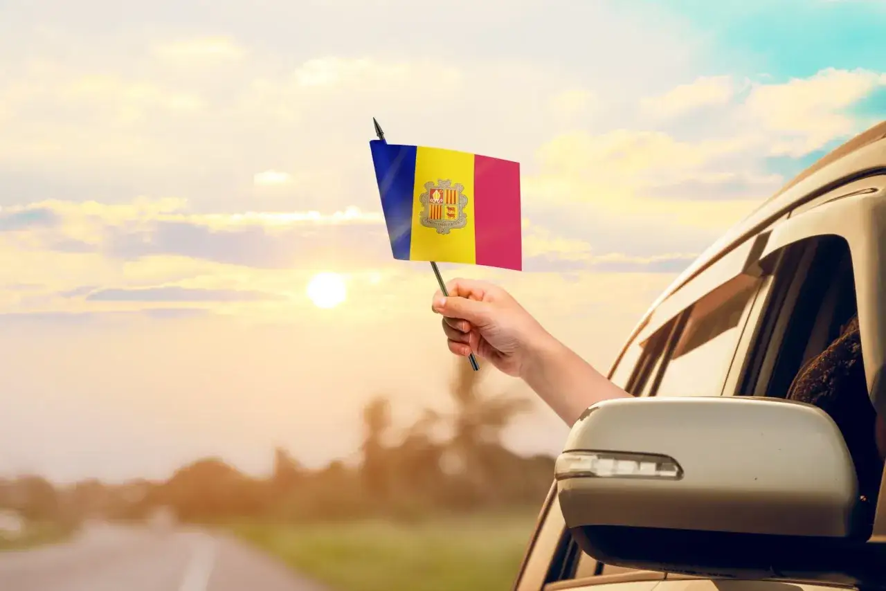 Ręka trzyma flagę Andory, kraju graniczącego z Francją i Hiszpanią, podczas podróży samochodem o zachodzie słońca.