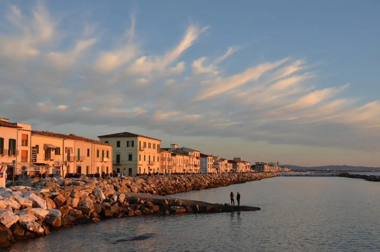 Marina di Pisa cosa vedere: case colorate sul lungomare, rocce e mare calmo al tramonto. Due persone osservano l'orizzonte.