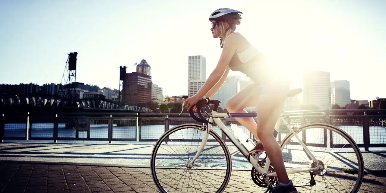 A female cyclist enjoys a sunny ride along a waterfront path, with city buildings in the background.