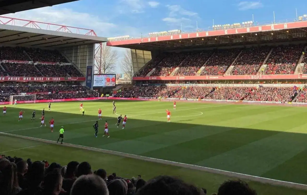 Mecz piłki nożnej na stadionie Nottingham Forest. Kibice w czerwonych barwach dopingują drużynę, a na boisku toczy się zacięta gra. Fani z pewnością śledzą najnowsze **Nottingham transfery**.