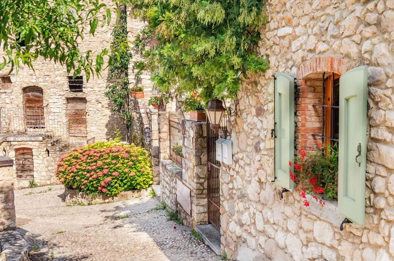 Un vicolo pittoresco sulla Gardesana Occidentale, con case in pietra, fiori rigogliosi e persiane verdi aperte.