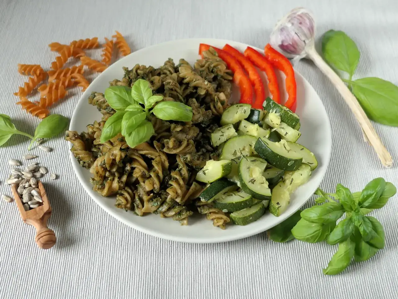 Makaron z aromatycznym pesto z bazylii, podany z cukinią, papryką i czosnkiem. Obok nasiona słonecznika i surowy makaron.