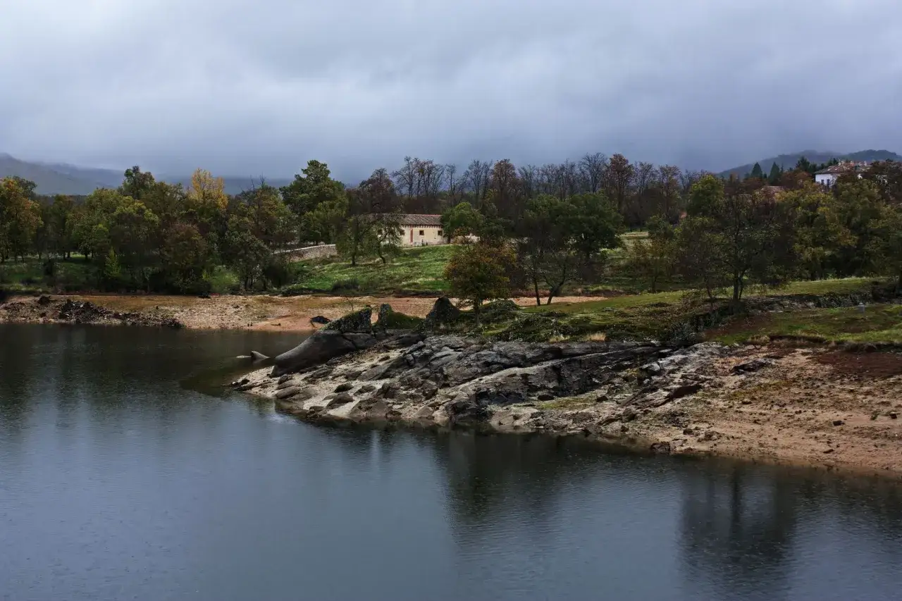 Vista del embalse del Pont&oacute;n con una casa en la ladera y &aacute;rboles oto&ntilde;ales bajo un cielo nublado.