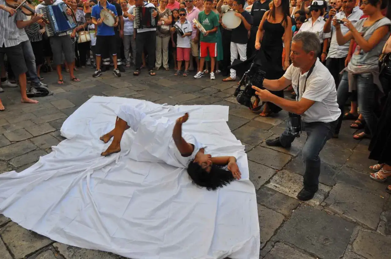 Danza e riprese video durante la festa patronale a Galatina. Una performer avvolta in un telo bianco si muove a terra, mentre un operatore filma la scena.