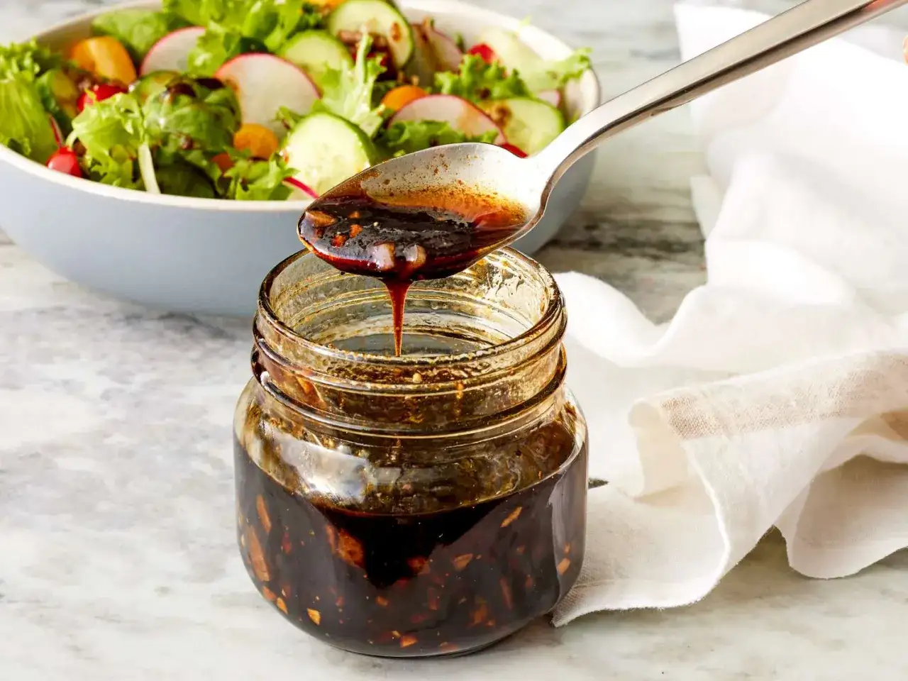 Łyżka polewa sałatkę gęstym, ciemnym sosem balsamicznym. Słoik z sosem stoi obok.