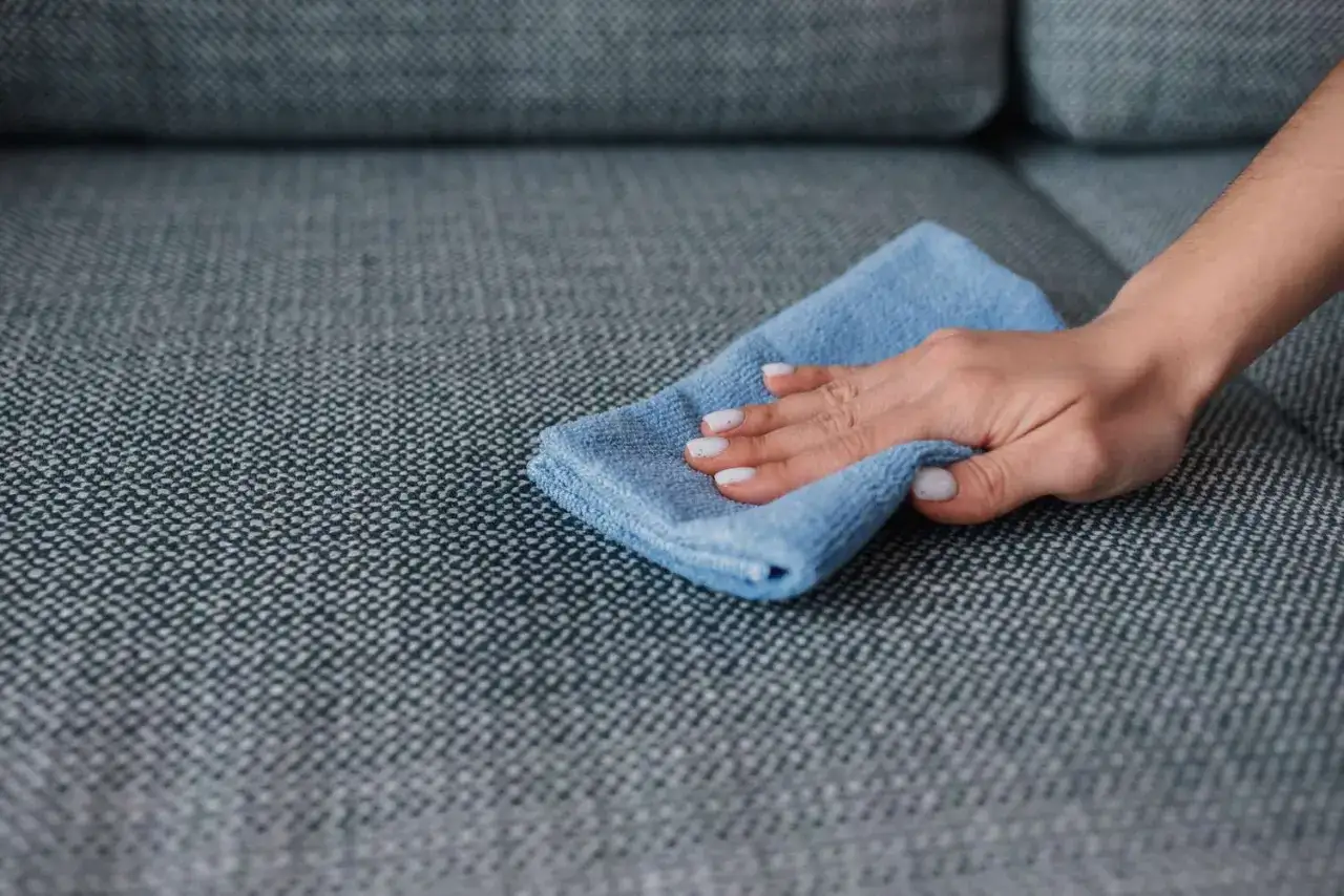 A hand with white nail polish gently wipes a grey couch with a blue microfiber cloth, demonstrating how to remove slime from couch.