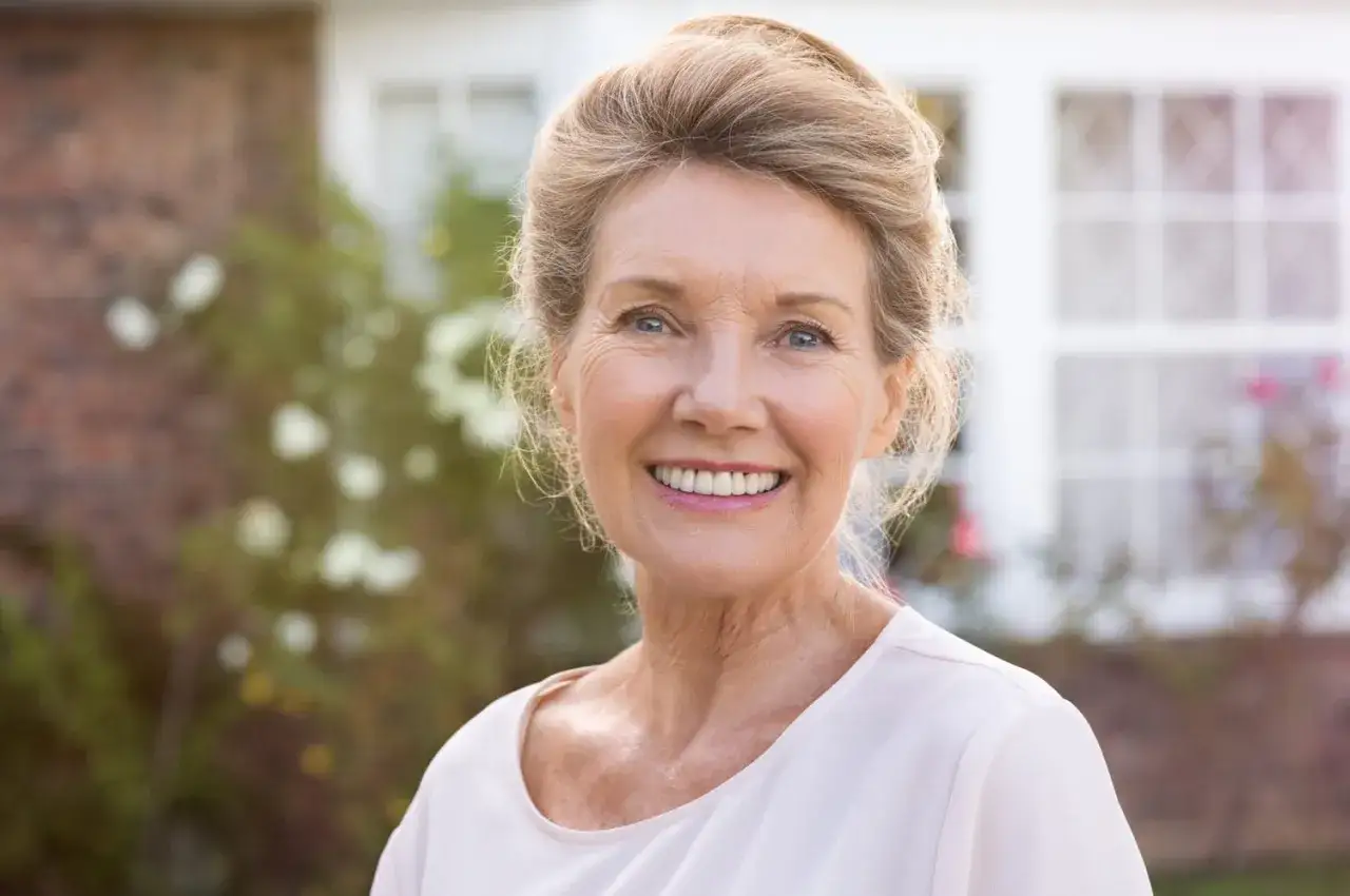Visage femme 80 ans souriant, cheveux blonds relevés, devant une maison.