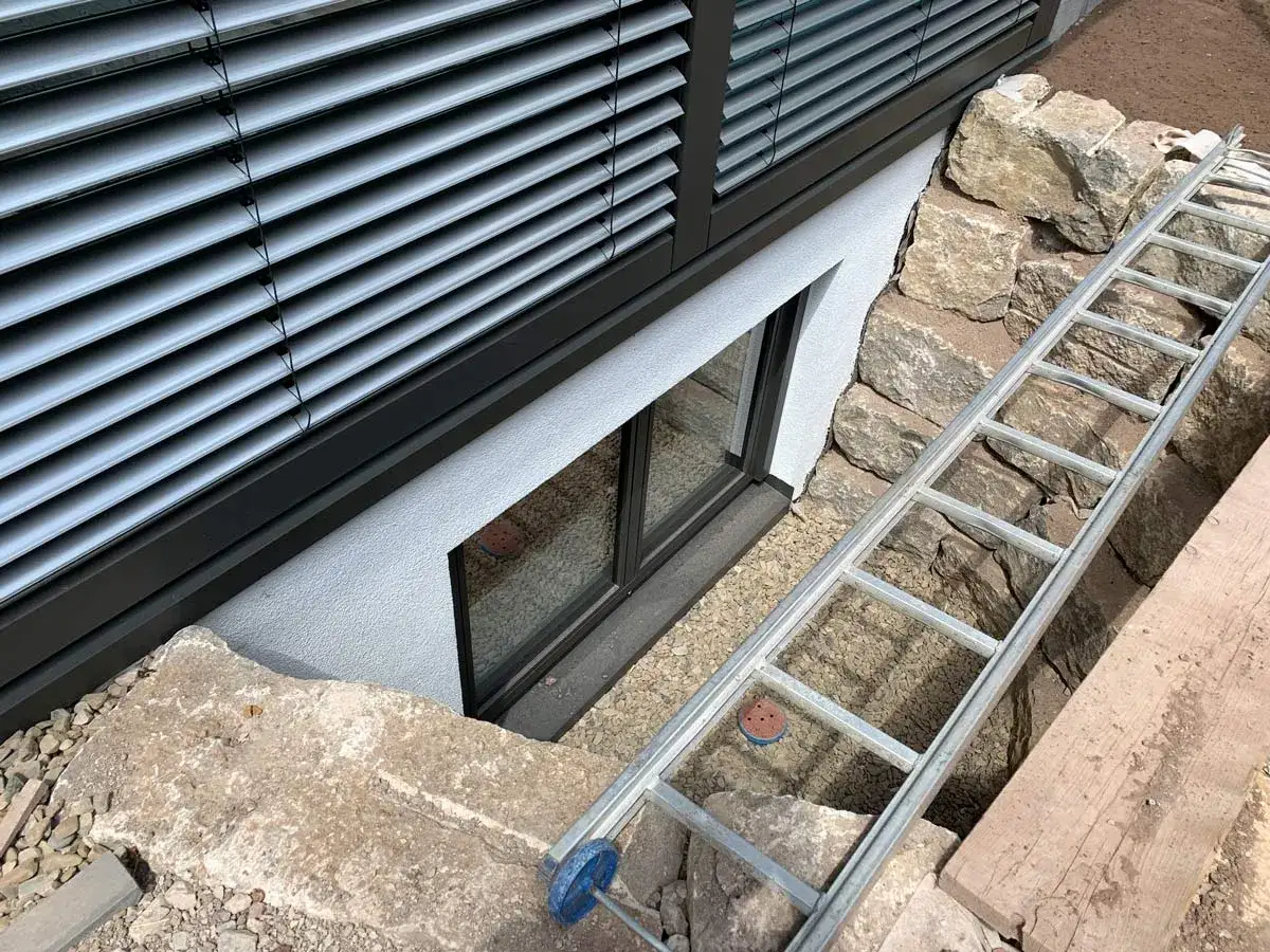 Eine Leiter liegt bereit, um das Kellerfenster auszutauschen. Gro&szlig;e Steine umgeben den Arbeitsbereich.