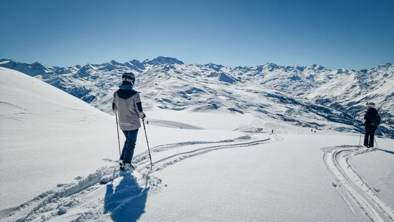 Vue panoramique d'une **randonnée Méribel hiver**. Deux skieurs contemplent un paysage montagneux enneigé sous un ciel bleu éclatant.