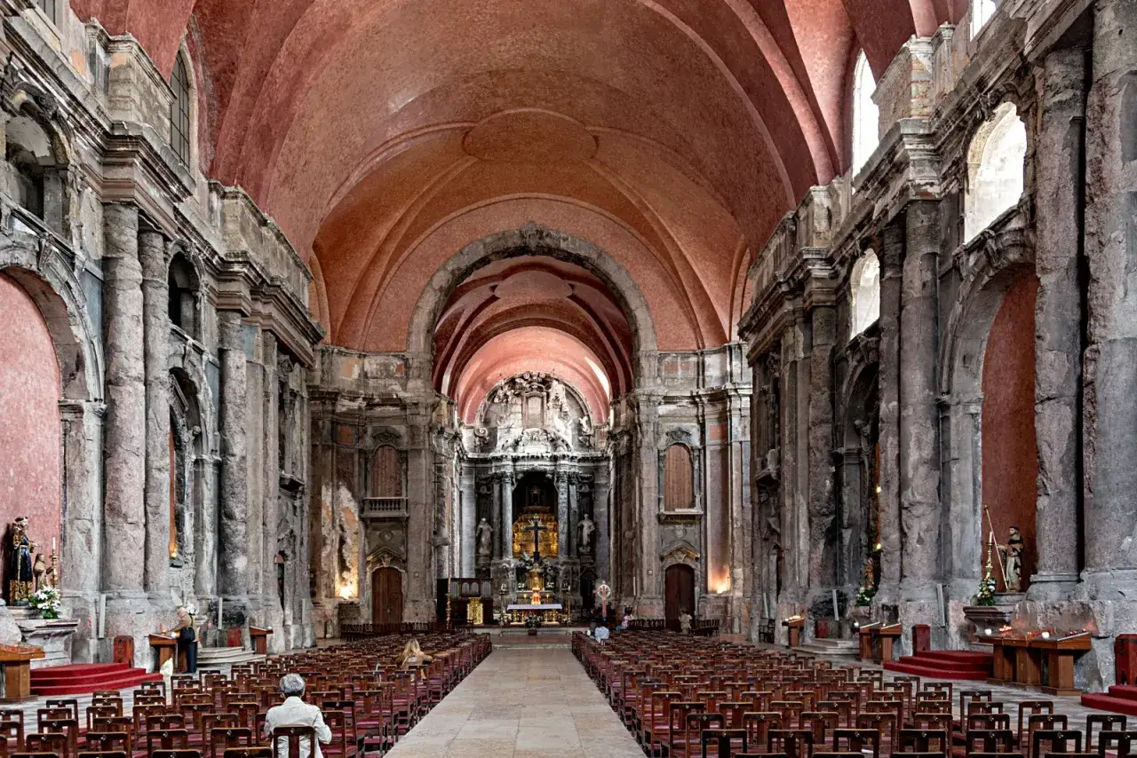 Das Innere einer beeindruckenden Kirche in Lissabon mit roten Gewölben und steinernen Säulen. Viele Holzbänke sind aufgestellt.